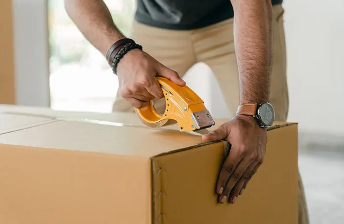 A person is holding a tape gun and cutting the cardboard box.